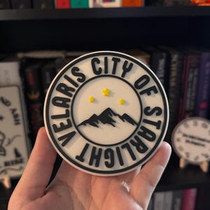 May include: A round, white coaster with a black border and text that reads "VELARIS CITY OF STARLIGHT". The center features a black mountain range and three yellow stars. A decorative item for home decor.