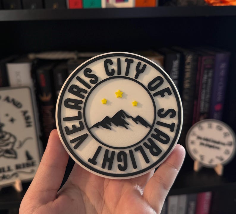 May include: A round, white coaster with a black border and text that reads "VELARIS CITY OF STARLIGHT". The center features a black mountain range and three yellow stars. A decorative item for home decor.