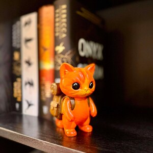 May include: A small, orange figurine of a cat with a backpack stands on a shelf. The cat has large, black eyes and a friendly expression. The backpack is tan. Books are in the background.