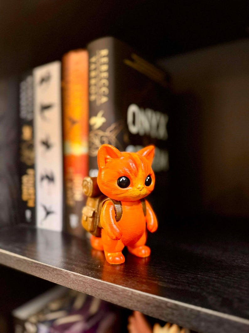 May include: A small, orange figurine of a cat with a backpack stands on a shelf. The cat has large, black eyes and a friendly expression. The backpack is tan. Books are in the background.