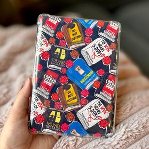 Pode incluir: Uma capa de telefone com um padrão repetido de capas de livros, incluindo títulos como "Locked Door" e "The Devil Wears Scrubs". A capa tem um fundo escuro com rosas vermelhas e é segurada em uma mão.