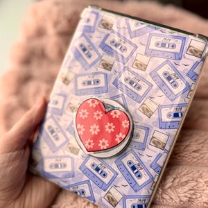 May include: A clear phone case featuring a pattern of light blue cassette tapes with the year "1984" printed on them. A red heart-shaped phone grip with white flower details is attached to the case.