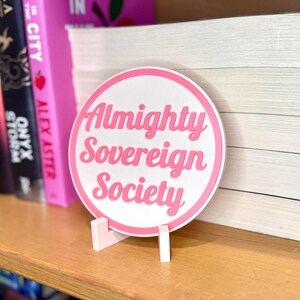 May include: A white round sticker with a pink border and the text "Almighty Sovereign Society" in pink lettering. The sticker is on a white stand and is sitting on a shelf with books.