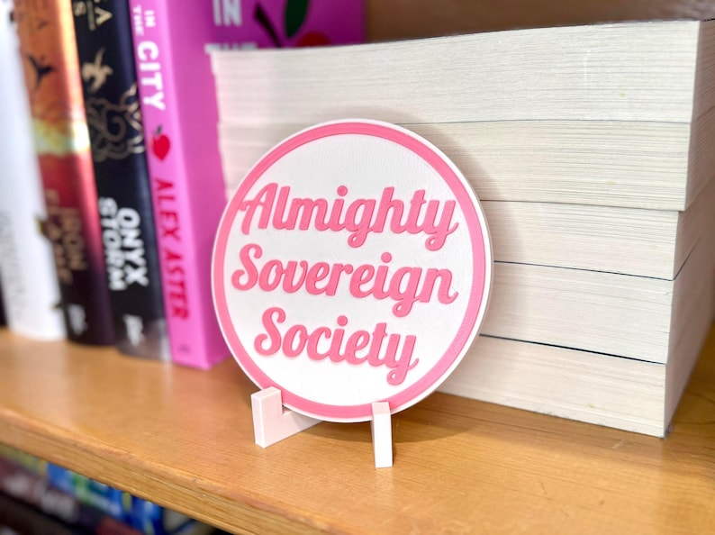 May include: A white round sticker with a pink border and the text "Almighty Sovereign Society" in pink lettering. The sticker is on a white stand and is sitting on a shelf with books.