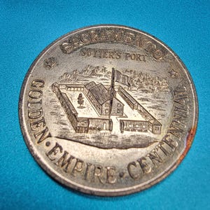 May include: A silver-colored commemorative coin with the text "SACRAMENTO SUTTER'S FORT" and "GOLDEN EMPIRE CENTENNIAL" encircling a detailed illustration of a fort. The coin is on a blue surface.