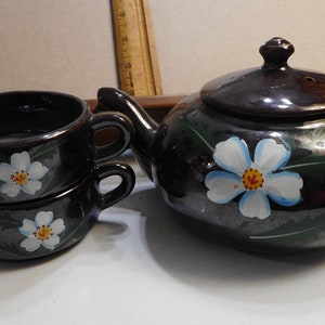 May include: A black tea set with white flowers painted on it. The set includes a teapot and two cups.