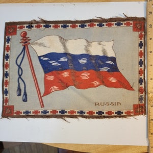 May include: A vintage fabric patch with a printed image of the Russian flag waving on a pole with tassels. The flag is white, blue, and red with a stylized design. The border of the patch is brown with a repeating pattern of red crosses and blue stars. The text "RUSSIA" is printed in brown at the bottom of the patch.
