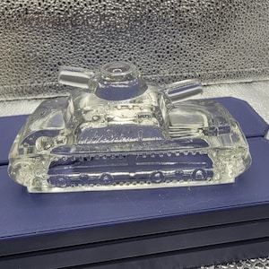 May include: A clear glass figurine shaped like a military tank. The object features a central turret, a protruding barrel, and molded details representing the tracks and wheels along the sides. The glass is transparent and reflects light.