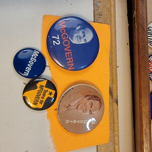 May include: Four round political campaign buttons featuring a man's face and the text "McGovern 72" and "Texans for McGovern".