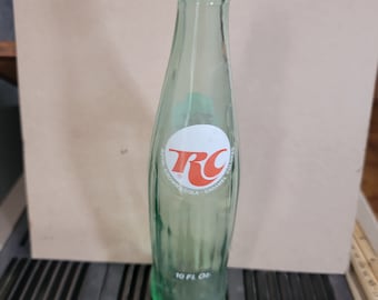 May include: A clear glass bottle with a green tint. The bottle has a white label with the text "Royal Crown Cola - Caramel Colored" and a red "RC" logo.
