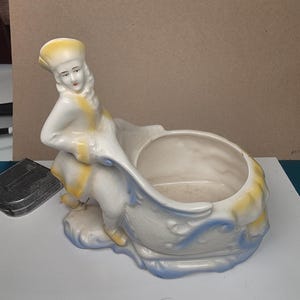 May include: A vintage ceramic planter featuring a woman in a flowing gown, leaning over a large, open bowl. The planter is primarily white with yellow and blue accents. The woman's hat and gown have yellow details, and blue swirls decorate the bowl.