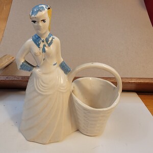 May include: A white ceramic figurine of a woman in a blue and white dress holding a white ceramic basket. The woman has blonde hair and blue eyes.