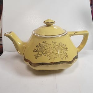 May include: A yellow ceramic teapot with a gold floral design and gold trim. The teapot has a lid with a small finial and a handle. The spout and handle are also trimmed in gold. A vintage tea set item.