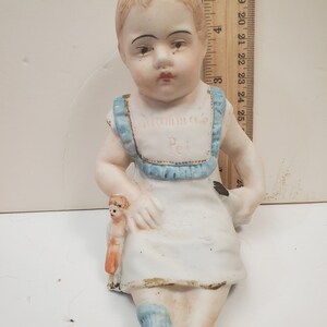 May include: A porcelain figurine of a child wearing a white dress with blue trim and a blue and white striped sock. The child is holding a smaller porcelain doll. The figurine has the words "Mammas Pet" written on the front.