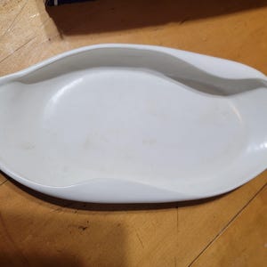 May include: A white, oval-shaped serving dish with a unique, curved design. The dish has a smooth, glossy finish and is set against a wooden surface. The dish is likely used for serving food.