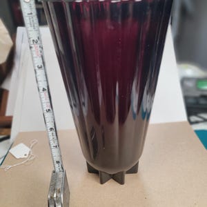 May include: A dark red glass vase with a fluted design, standing approximately 8 inches tall. The vase has a star-shaped base and a flared top. A metal tape measure is next to the vase.