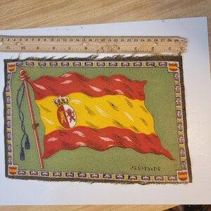 May include: A vintage fabric patch with a green background and a red and yellow Spanish flag with a coat of arms. The flag is waving and the patch has a decorative border. The text "SPAIN" is printed in black at the bottom of the patch.