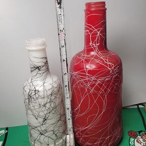 May include: Two glass bottles standing next to a metal measuring tape. The smaller bottle is white with black scribbled lines, and the larger bottle is red with white scribbled lines. The measuring tape shows the red bottle is 10 inches tall.