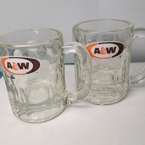 May include: Two clear glass mugs with thick handles and a dimpled base design. Each mug features the A&W logo printed on the front, consisting of an orange oval border with the letters A&W in white and brown text against a dark brown background.