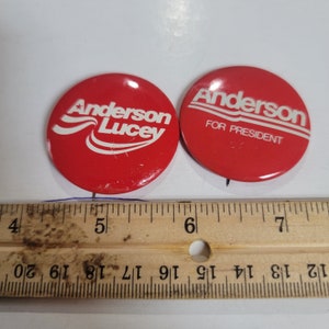 May include: Two red and white political campaign buttons. The left button says "Anderson Lucey" with a wavy line and star. The right button says "Anderson for President" with a wavy line.