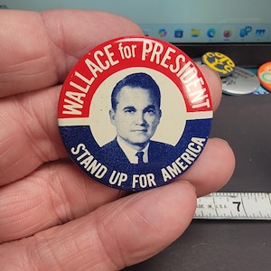 May include: A vintage political campaign button for Wallace for President. The button features a portrait of a man in a suit and tie, with the text "Wallace for President" in red and "Stand Up For America" in blue.