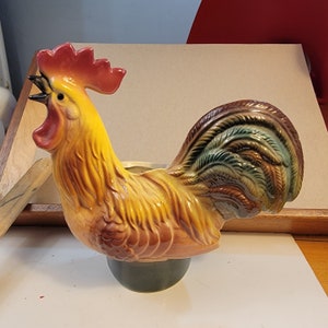 May include: A ceramic rooster figurine with a bright red comb and wattles, brown and yellow feathers, and a green and brown tail. The rooster is standing on a green base.