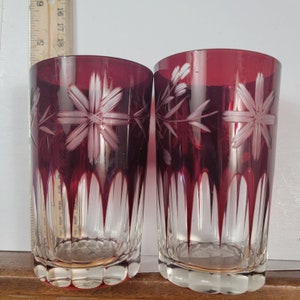 May include: Two ruby red glass tumblers with etched floral designs. The glasses have a clear base and a ribbed design around the bottom.