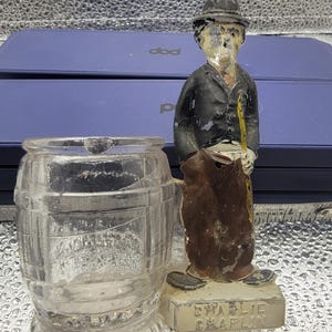 May include: A clear glass barrel-shaped container next to a painted metal figurine of Charlie Chaplin wearing a black hat, black jacket, and brown trousers, holding a yellow cane. The base of the figurine is inscribed with the name Charlie Chaplin.
