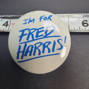 May include: A vintage campaign button with the text "I'm FOR FRED HARRIS!" in blue lettering on a white background. The button is round and appears to be made of metal. A ruler is in the background.