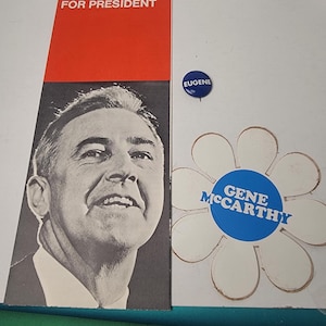 May include: A political campaign set including a rectangular poster with a black and white portrait of McCarthy and the text McCarthy FOR PRESIDENT, a small blue circular pin with the text EUGENE, and a white flower-shaped sticker with a blue center reading GENE McCARTHY, shown with a measuring tape indicating a width of approximately 6 inches.