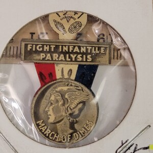 May include: A vintage March of Dimes pin with a gold-colored Walking Liberty half dollar design. The pin features a red, white, and blue ribbon with the text "FIGHT INFANTILE PARALYSIS".