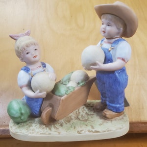 May include: A porcelain figurine of two children, a boy and a girl, dressed in overalls, pushing a wheelbarrow filled with vegetables. The boy is wearing a cowboy hat and holding a white melon. The girl is holding a white melon and has a pink bow in her hair.