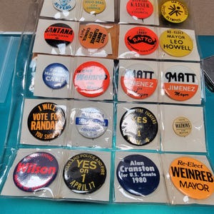 May include: A collection of vintage political campaign buttons in a clear plastic sheet. The buttons feature various names, slogans, and logos, including "Re-Elect Weinreb Mayor" and "Yes on 1 April 17". The buttons are in various colors.