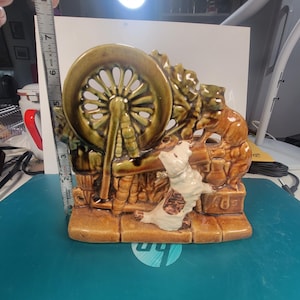 May include: A ceramic figurine depicting a spinning wheel with a dog and cat. The figurine is primarily brown and green, with the dog in white and brown. The figurine measures approximately 5 inches in height.