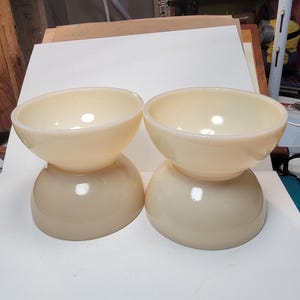 May include: Two vintage, cream-colored glass bowls. Each bowl has a rounded base and a flared, open top. The bowls are stacked, with the top bowl resting on a smaller, inverted bowl. The glass has a glossy finish.