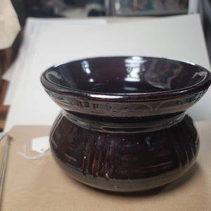 May include: A dark brown ceramic bowl with a glossy finish. The bowl has a wide, shallow top section and a rounded, textured base. The interior of the bowl is a deep, rich brown color. The bowl is likely handmade and suitable for serving or display.
