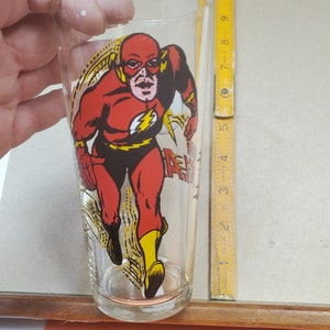 May include: A clear glass featuring a full-body image of The Flash in his classic red and yellow costume. The superhero is depicted in a running pose with the word "Flash" printed on the side. The glass is held by a hand.