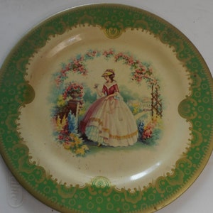 May include: A vintage decorative plate with a green and gold border. The center of the plate features a woman in a white dress standing in a garden setting.