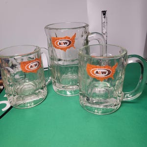 May include: Three clear glass mugs with handles, each featuring an orange map of the United States and the A&W logo. A measuring tape is visible behind the center mug, showing a height of approximately 5 inches for the tallest glass vessel.