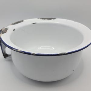 May include: White enamelware chamber pot with a blue rim and handle. The pot has some wear and tear, with chipped paint around the rim. The interior is white and clean, and the pot has a rounded shape.