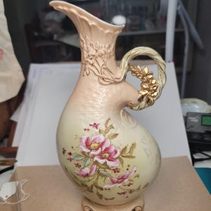 May include: A decorative, vintage-style pitcher with a cream and peach color scheme. The pitcher features a floral design with pink flowers and green leaves. The handle is adorned with gold accents, and the base has an ornate design.