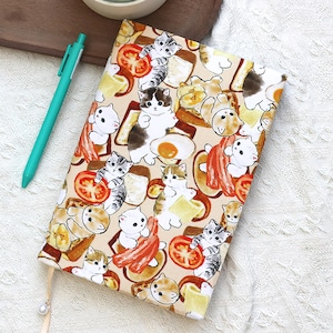 May include: A beige notebook with a repeating pattern of cartoon cats, bacon, eggs, tomatoes, and toast. A teal pen rests beside the notebook. A cream-colored ribbon with a pearl charm is attached to the notebook.