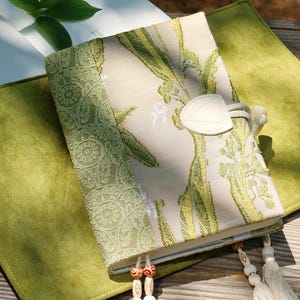 May include: A decorative journal with a cream-colored cover featuring green floral embroidery and a leaf-shaped closure. The journal rests on a green suede-like surface, with a matching green patterned border. Tassels and wooden beads hang from the journal's spine.