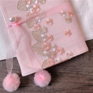 May include: A light pink, rectangular clutch with floral embroidery and a pearl clasp. The clutch features a sheer ribbon and two fluffy pink pom-poms. The design includes embroidered flowers and leaves.