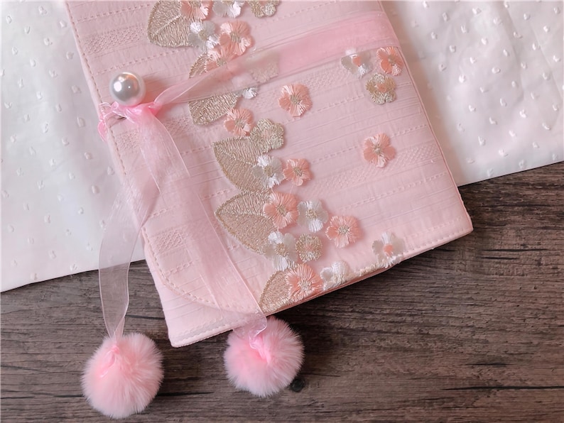 May include: A light pink, rectangular clutch with floral embroidery and a pearl clasp. The clutch features a sheer ribbon and two fluffy pink pom-poms. The design includes embroidered flowers and leaves.