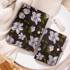 Pode incluir: Dois cadernos pretos com um padrão floral de flores brancas e folhas verdes. Os cadernos têm um aspeto texturizado e são presos com um botão dourado e um fecho de corda preta. Um dos cadernos é maior do que o outro.
