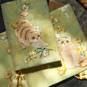Puede incluir: Tela verde claro bordada con imágenes detalladas de gatos persas y acentos florales en amarillo y blanco. Los gatos están representados en varias poses, con costuras y sombreados intrincados. La tela tiene una apariencia texturizada con motas doradas.