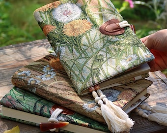 May include: A stack of floral-patterned journals with a tapestry-like design. The covers feature dandelions and other wildflowers in shades of green, brown, and orange. Each journal has a unique closure, including a leather tab and a beaded tassel.