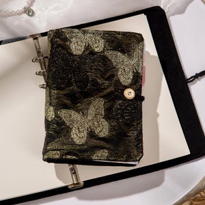 May include: A black journal with a gold butterfly pattern. The journal has a button closure and is open to blank pages. The journal is sitting on a white surface.