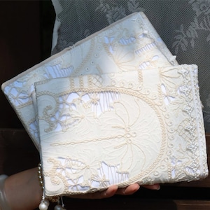 May include: Two rectangular notebooks featuring cream-colored lace covers with intricate floral and geometric embroidery patterns. The lace overlays a solid white base, and the spines are finished with a decorative white lace trim border.